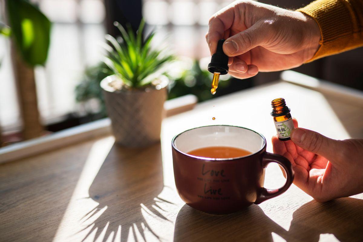 A person adding a drop of essential oil to a cup of tea, with sunlight streaming in and a potted plant in the background.