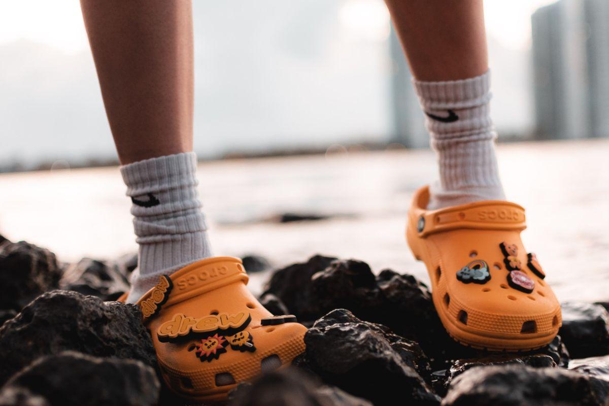 Person wearing bright orange Crocs decorated with charms, standing on rocks near the water.