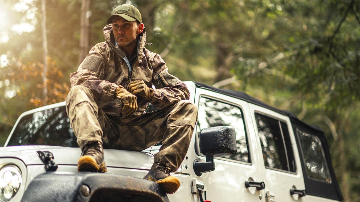 Man in camo gear sitting on a white off-road vehicle in the woods, showing a typical setup for outdoor hunting with different types of guns.