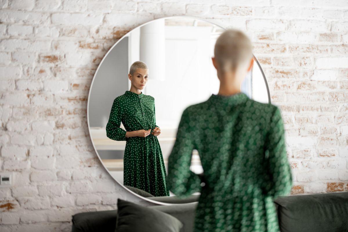 A woman in a green polka dot dress looking at her reflection in a round mirror mounted on a brick wall.