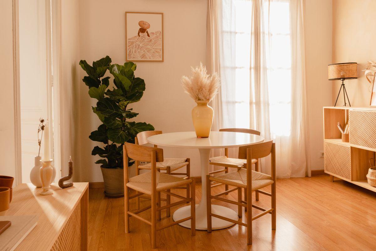 A cozy, minimalist dining room with a round table, wooden chairs, potted plant, and soft natural light filtering through sheer curtains.