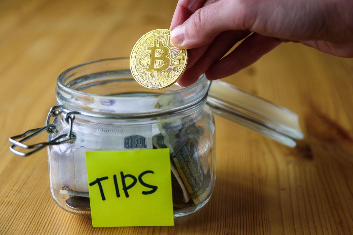 Hand placing a Bitcoin coin into a glass tip jar labeled 'TIPS' filled with cash.
