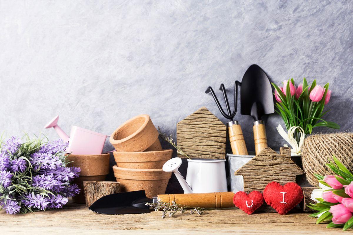 Gardening tools and decor on a wooden surface, including terracotta pots, small shovels, a pink watering can, twine, and flowers, with two red heart-shaped decorations in the foreground.
