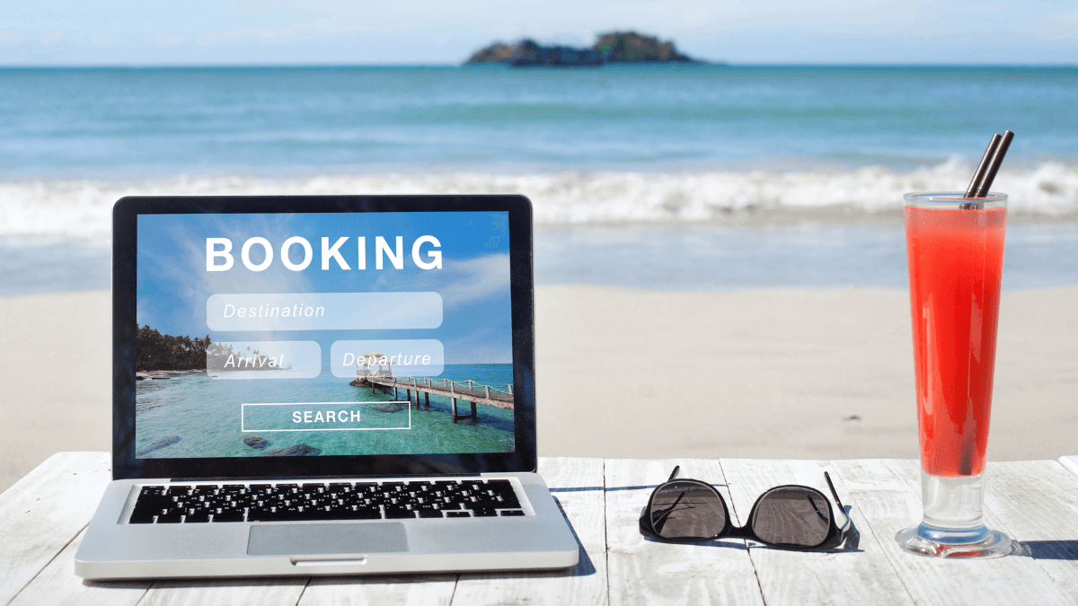 Online travel booking on a laptop at a beachside setting with sunglasses and a refreshing drink, showcasing a tropical getaway planning experience.