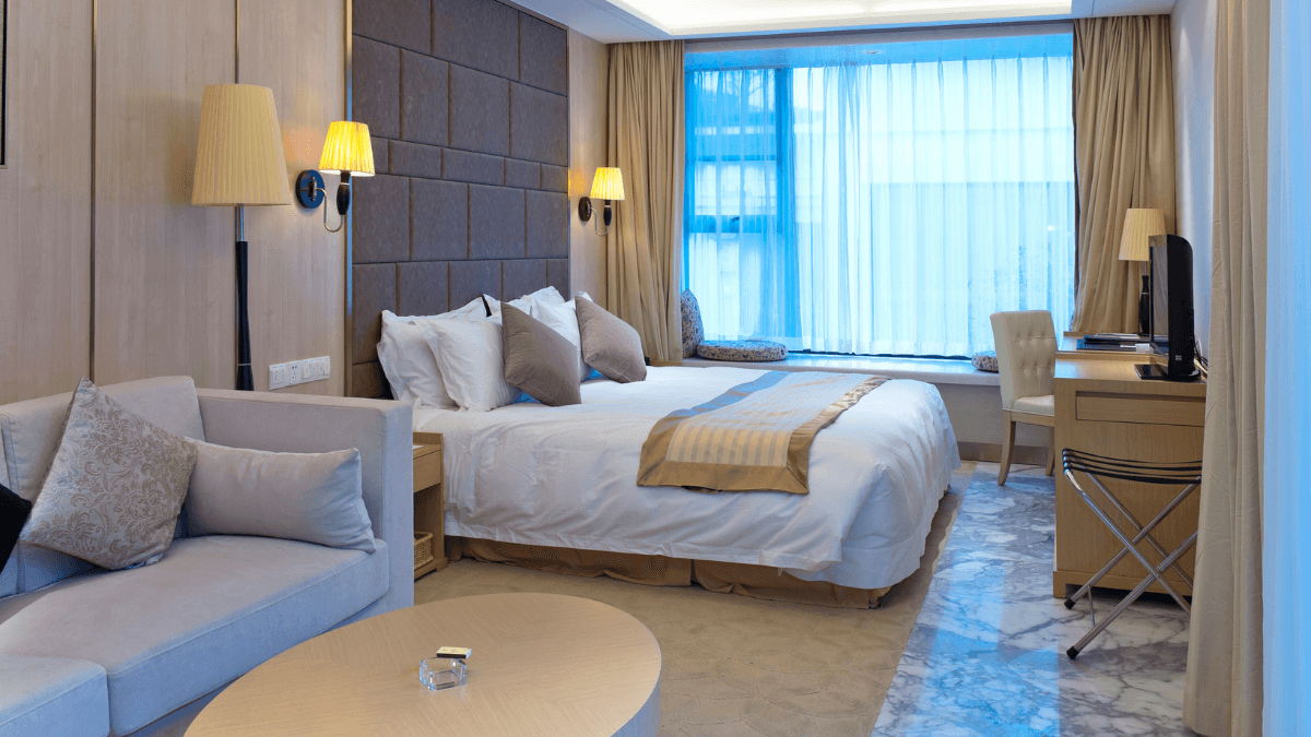 Modern hotel room interior featuring a cozy double bed with decorative pillows, elegant wall lamps, a stylish armchair, and a well-lit window with sheer curtains, creating an inviting and relaxing atmosphere.