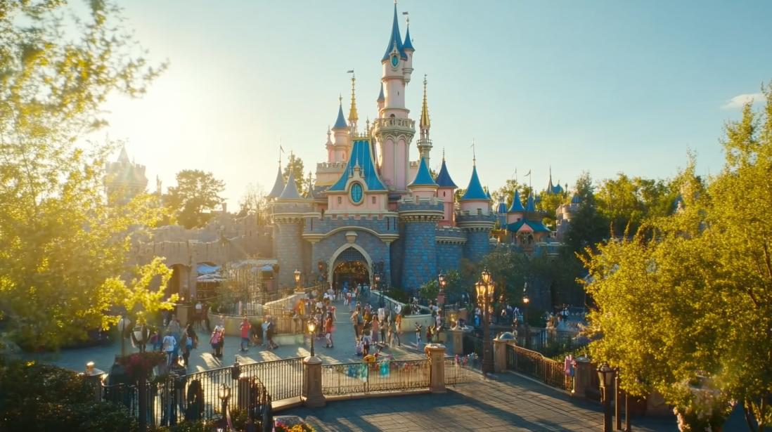 A beautifully lit Disneyland park at sunset, with families walking toward a grand Victorian-style building in the distance. The park features lush gardens, glowing streetlights, and lively crowds enjoying the magical atmosphere. Ideal for travel, family vacations, or theme park experiences.