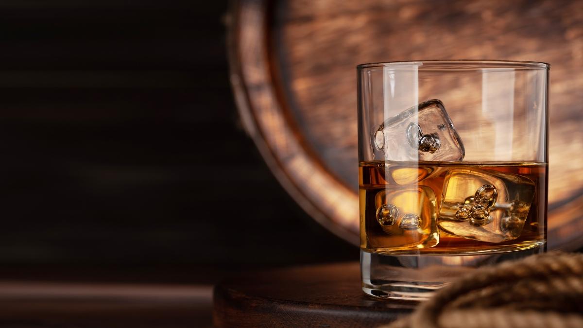 A glass of whiskey with ice sits on a wooden surface in front of a barrel, perfect for showcasing the rich color of Son of a Peat whiskey.