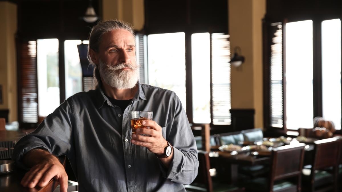 Man with a gray beard holding a glass of whiskey in a restaurant, showing a relaxed moment that pairs well with sipping premium spirits.