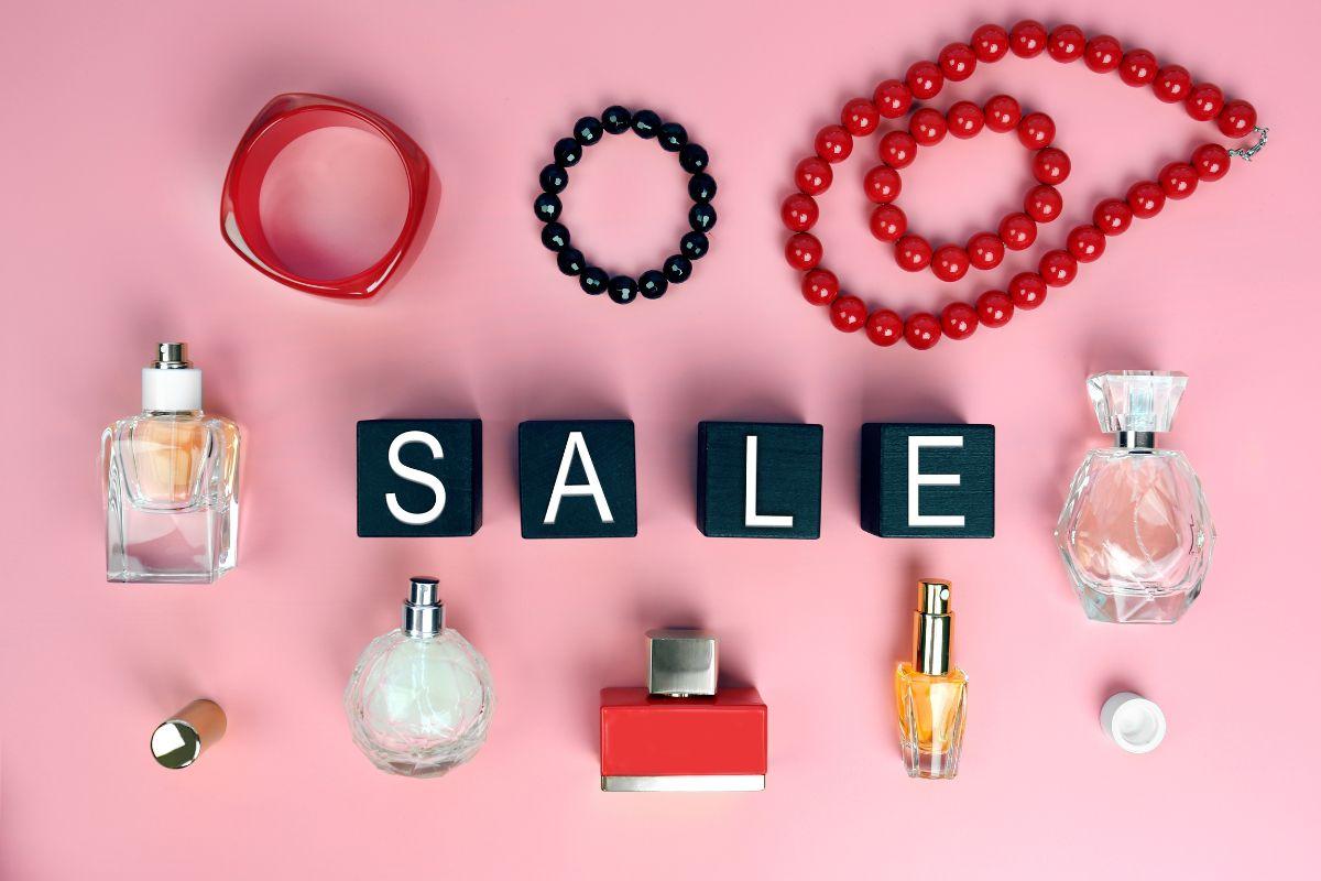 Perfume bottles, bracelets, and a necklace arranged around black blocks spelling 'SALE' on a pink background.