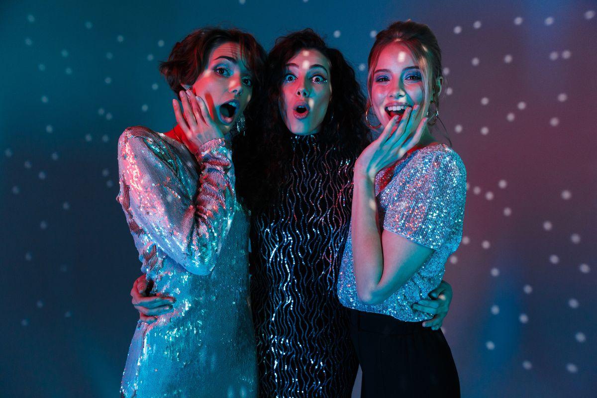 Three women in sparkly outfits posing with surprised expressions under disco lights, creating a festive party atmosphere.