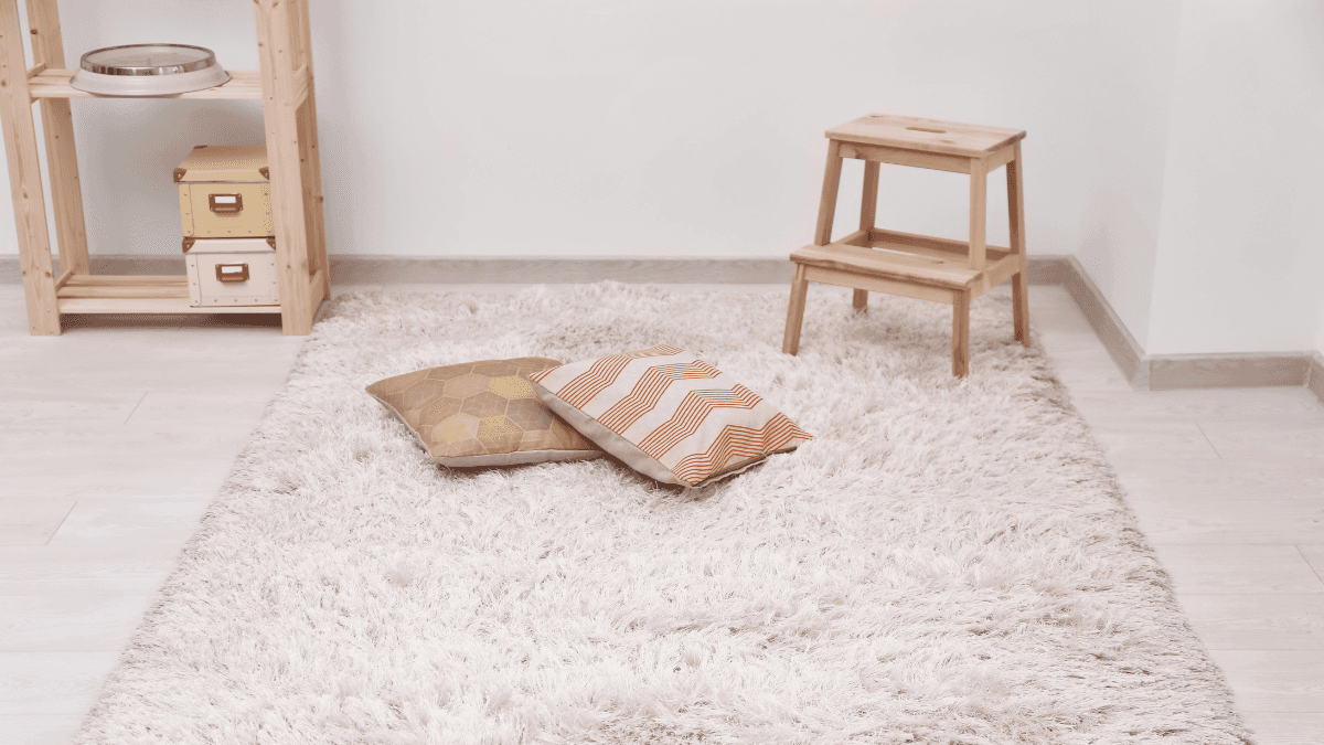 Cozy room with a soft, fluffy beige rug, two decorative pillows, a wooden step stool, and storage shelves in the background.
