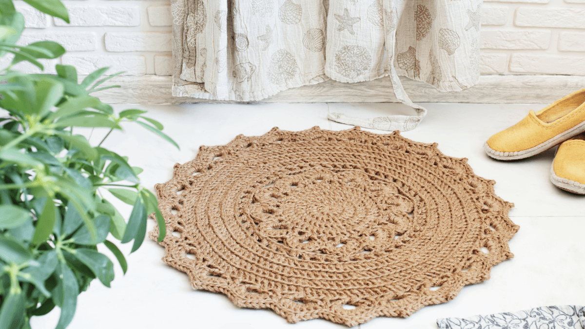 Round crochet rug in a natural brown tone placed on a light floor, accompanied by yellow shoes and leafy green plant, adding a cozy and rustic charm to the space.