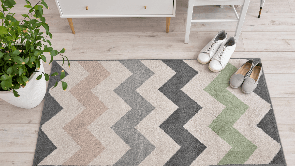 Cozy, chevron-patterned rug with pastel and neutral tones, placed beside a small chair and cabinet, complete with a touch of greenery and neatly positioned shoes for a warm, welcoming entryway.