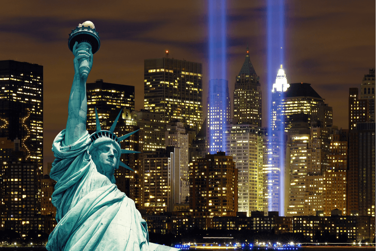 The Statue of Liberty in the foreground with the New York City skyline at night and the Tribute in Light beams shining in the background.