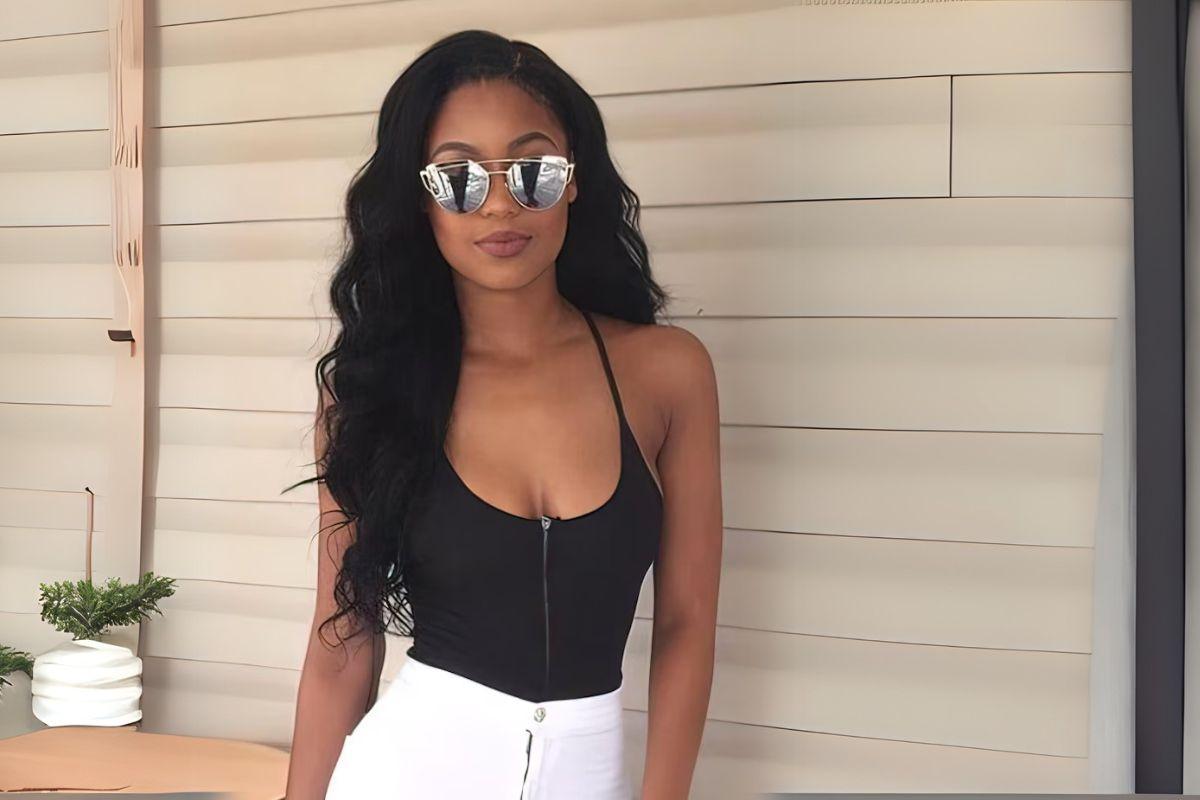 Woman with long, wavy black hair wearing sunglasses, a black halter top, and white pants, standing against a paneled wall.