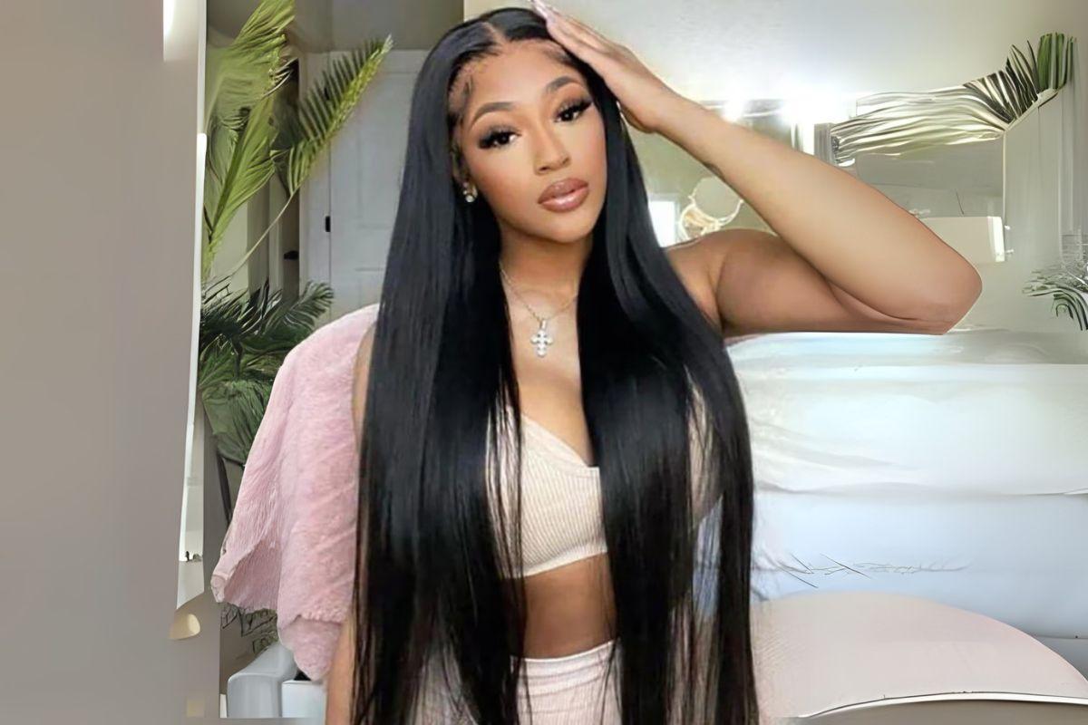 Woman with long, straight black hair wearing a light-colored top, posing with one hand on her head in a room with modern decor and greenery.