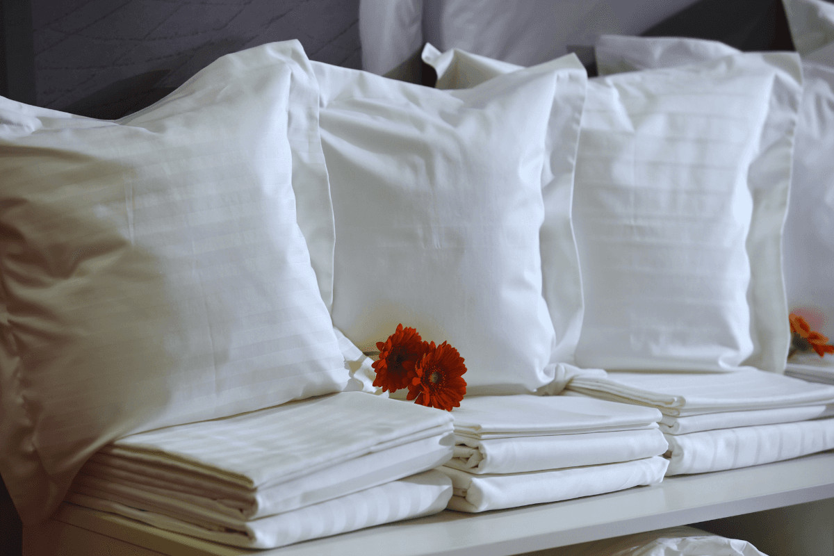 Neatly stacked white pillows and bed linens with orange flowers placed as accents on a display shelf.