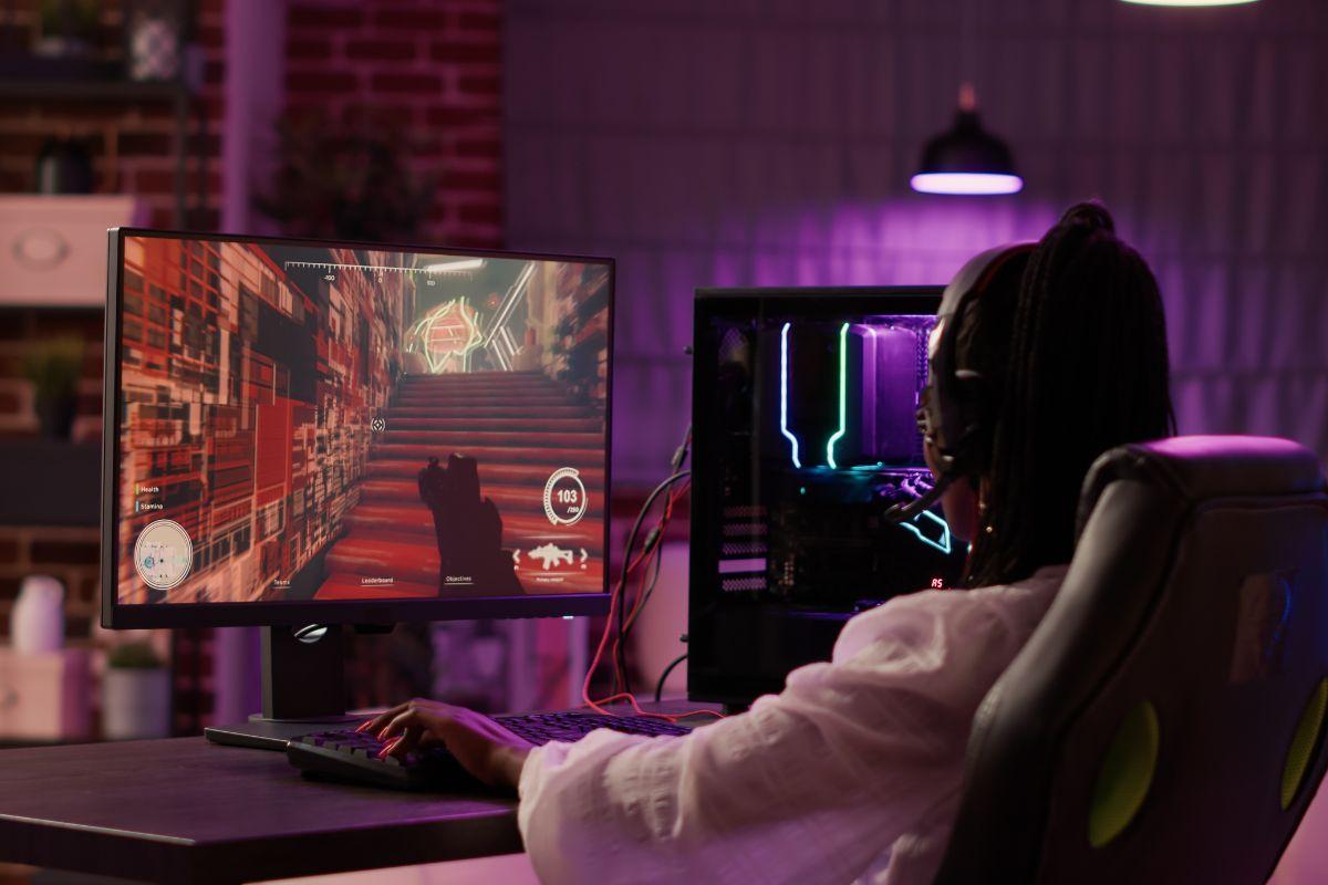 Gamer wearing a headset, playing a first-person shooter game on a monitor, with a gaming PC featuring colorful LED lights in a cozy, dimly lit room.