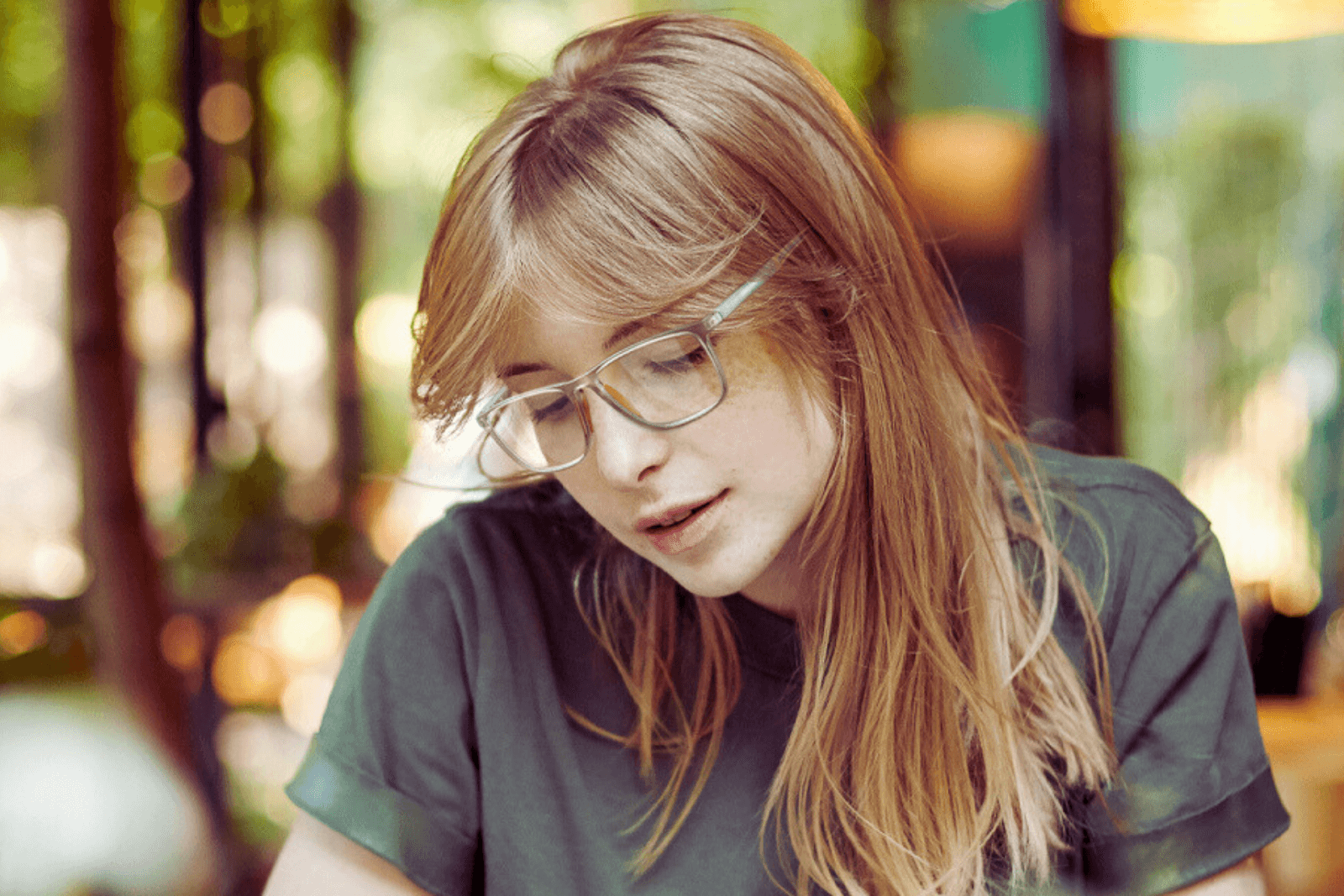 A young woman with long blonde hair and clear-framed glasses looking down, wearing a green shirt, in a softly lit outdoor setting with blurred background greenery.