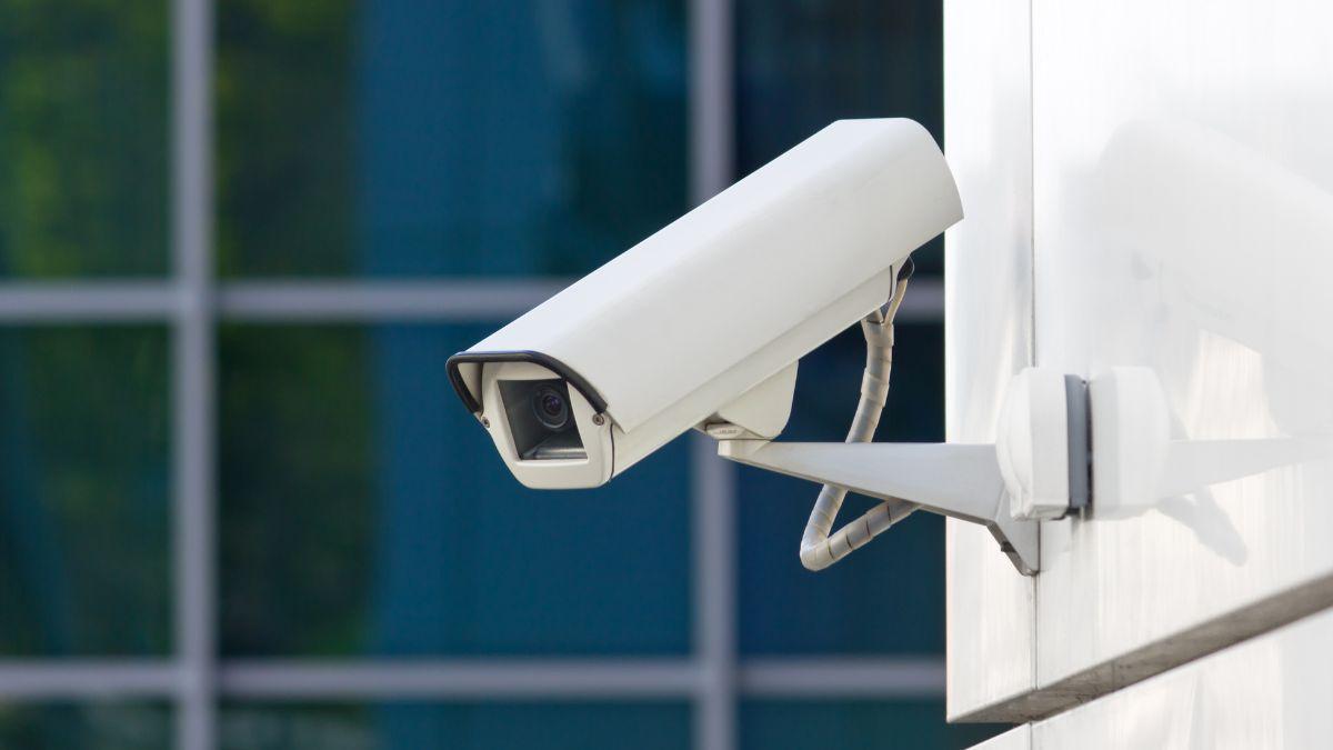 A white security camera is mounted on a wall, overlooking an area with glass windows in the background.