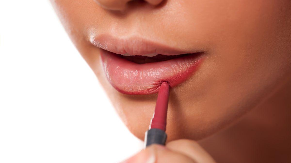 A close-up of a woman applying pinkish-red lip liner to her lower lip for a defined and polished look.