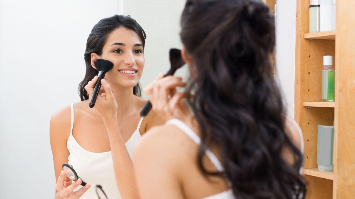 A woman with long dark hair smiles while applying makeup with a brush in front of a mirror.