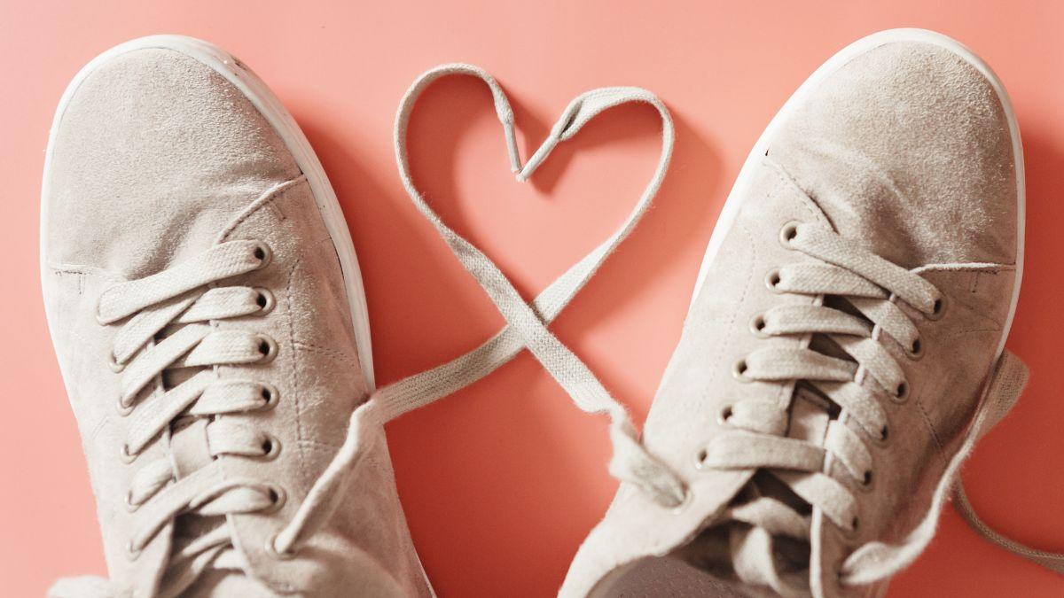 A pair of beige sneakers on a pink background with their laces shaped into a heart.