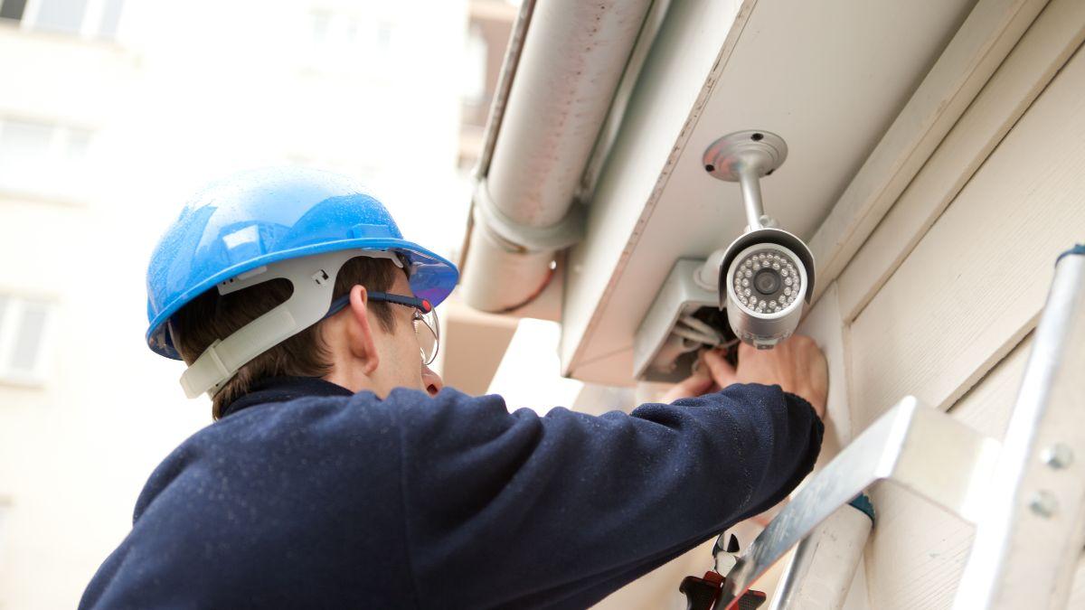 A worker in a blue hard hat installs a security camera on the side of a building.