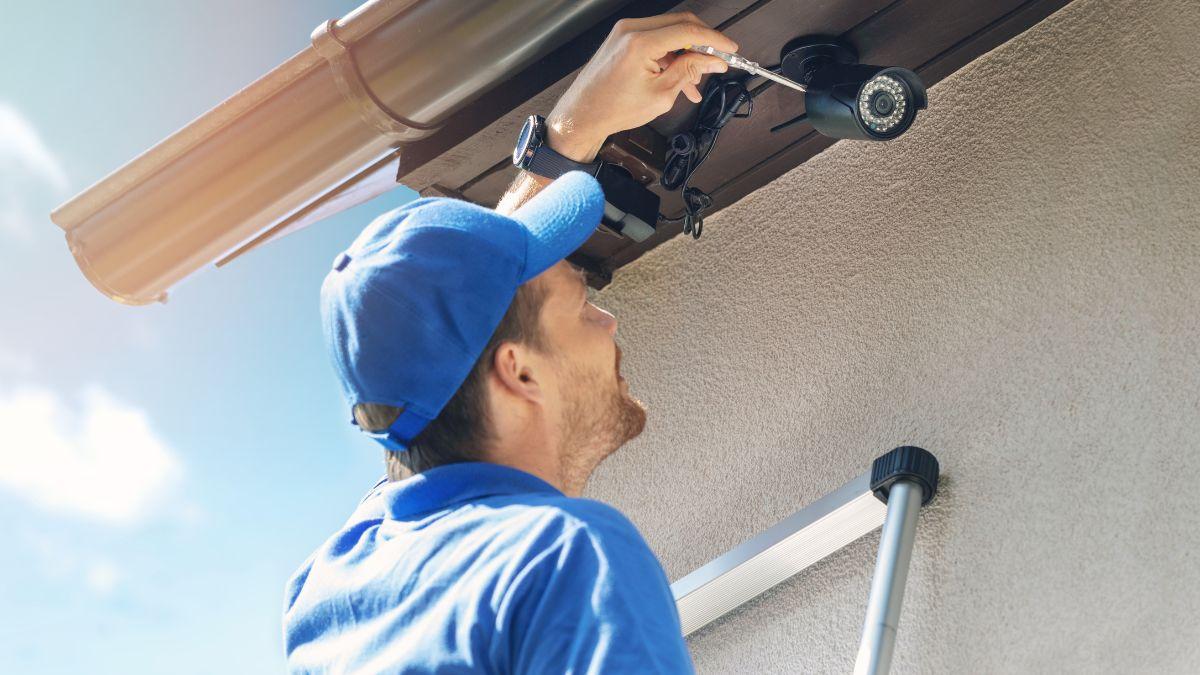 A worker in a blue uniform and cap is installing a security camera under the roof using a screwdriver.