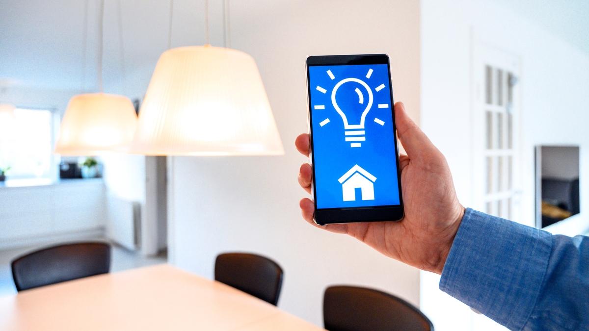 A person holds a smartphone displaying a smart home lighting app, controlling hanging lights in a modern dining area.