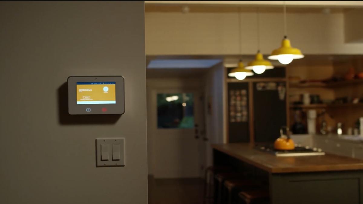A smart home control panel is mounted on a wall, displaying a lit screen, with a modern kitchen in the background featuring hanging yellow pendant lights.