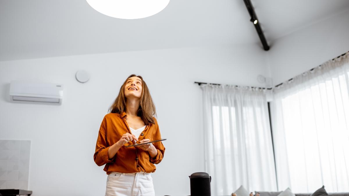 A woman in a modern, bright living room controls smart lighting using a tablet, looking up at the ceiling light with a smile.