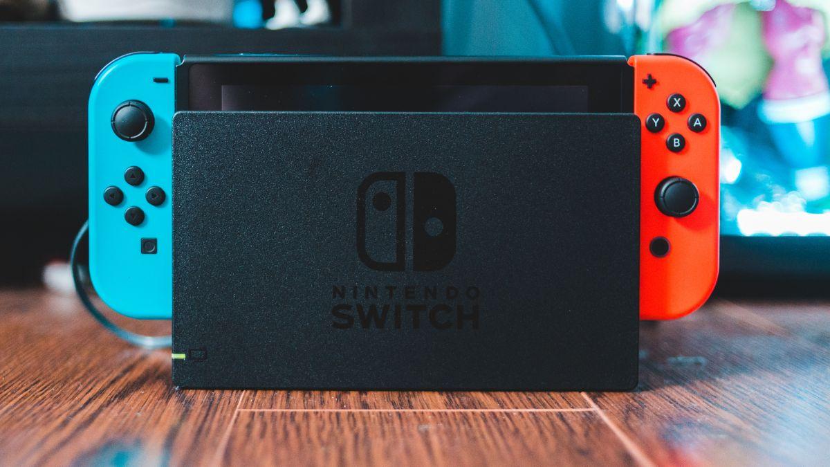 A Nintendo Switch console with blue and red Joy-Con controllers sits in its dock on a wooden floor.
