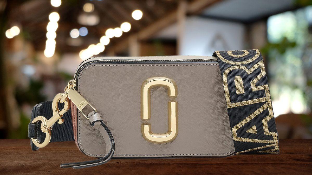 A small, gray designer handbag with a gold logo and a thick black strap with gold lettering sits on a wooden surface.