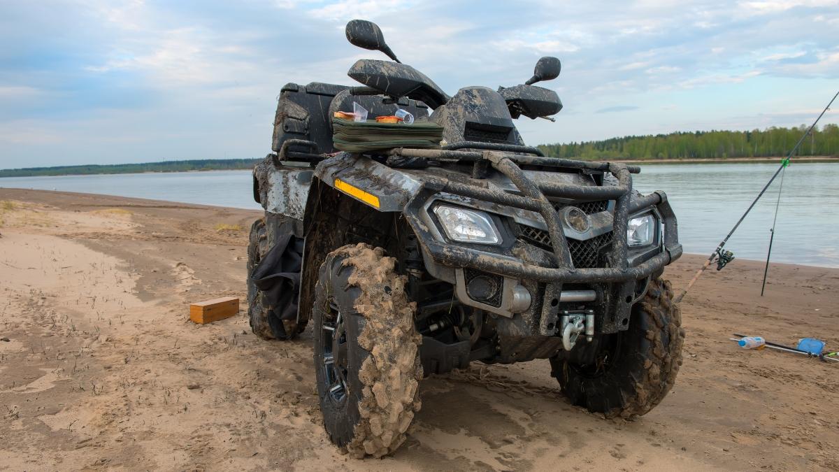 Mud-covered ATV parked on a sandy riverbank showing rugged tires and outdoor off-road use.