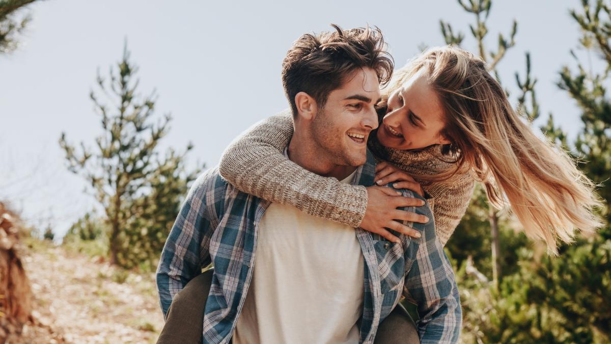Smiling couple enjoying a piggyback ride outdoors surrounded by pine trees, symbolizing intimacy and connection for a Hims Sex Ultra Thin Condoms lifestyle concept.