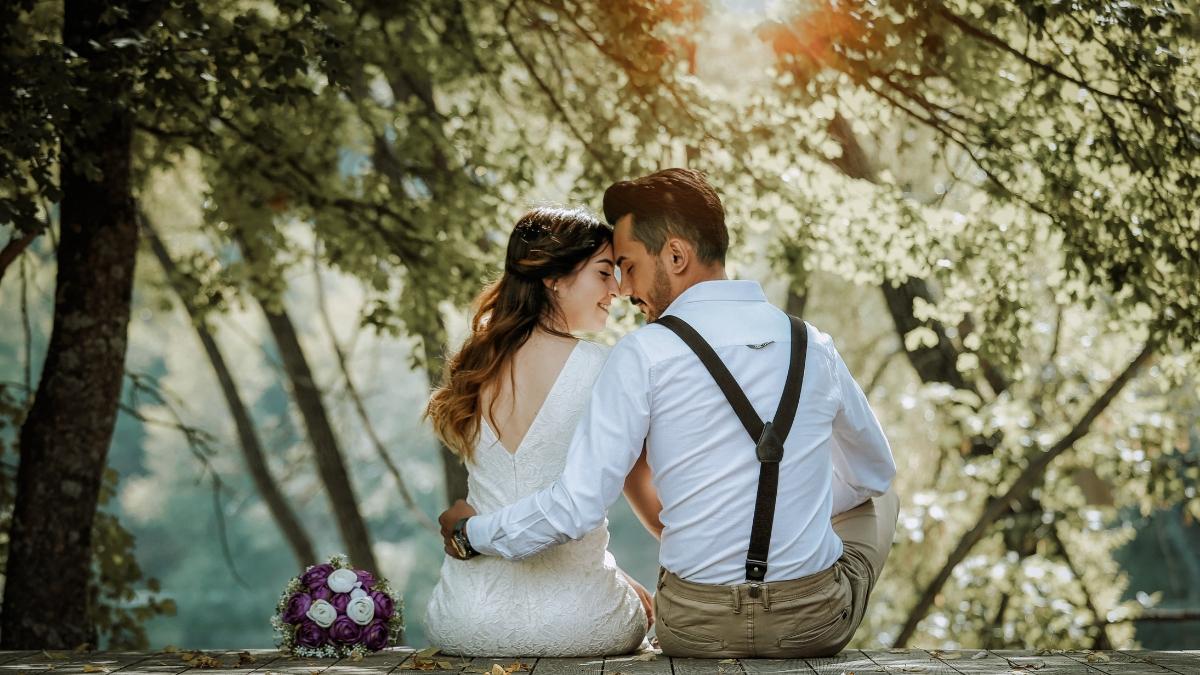 A newlywed couple sitting close together on a wooden deck surrounded by trees, sharing an intimate moment outdoors after their wedding, with a purple bridal bouquet placed beside them.