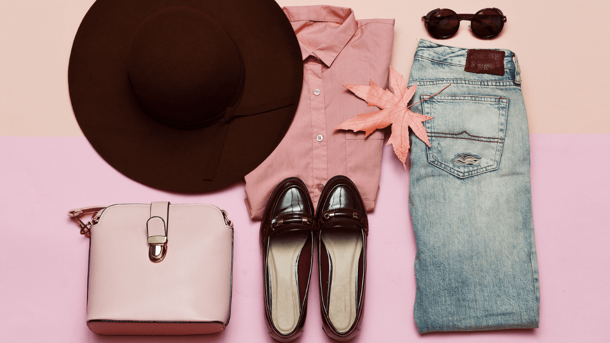 A flat lay of stylish autumn essentials including a wide-brimmed brown hat, a pink blouse, dark loafers, blue jeans, sunglasses, a soft pink handbag, and a fall leaf for a seasonal touch.