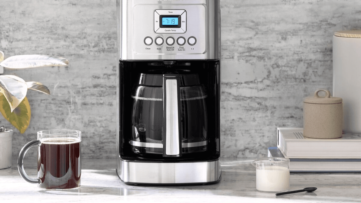 A sleek drip coffee maker with digital display on a kitchen counter, accompanied by a steaming cup of coffee, milk, and a small jar, set against a textured stone wall backdrop.