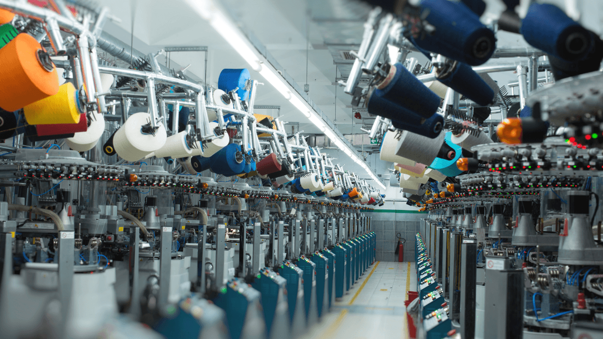 An industrial textile manufacturing plant with large spools of colored thread suspended above automated machinery, under bright overhead lighting in a spacious, modern facility.