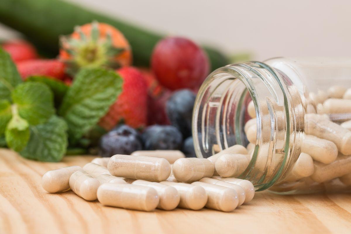 An open glass jar with supplement capsules spilling out, surrounded by fresh fruits and herbs on a wooden surface.