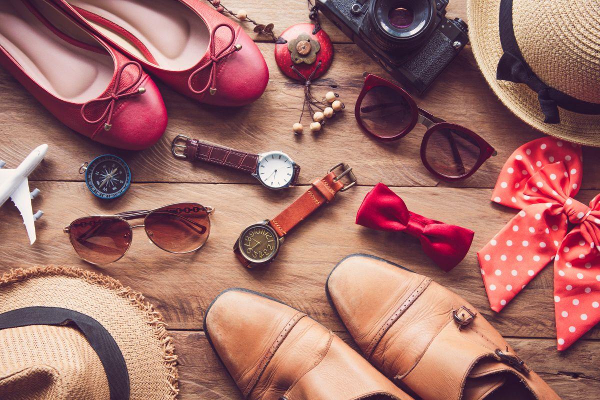 A collection of travel accessories on a wooden surface, including red shoes, sunglasses, watches, hats, a camera, and a red bow tie.