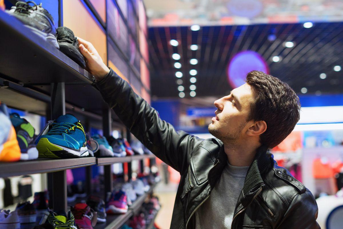 A man in a leather jacket browsing sneakers on a store shelf, examining a pair closely with various athletic shoes displayed around him.