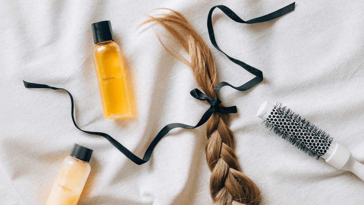Haircare essentials including shampoo, conditioner, a hairbrush, and a braided lock of hair tied with a black ribbon on a soft surface.