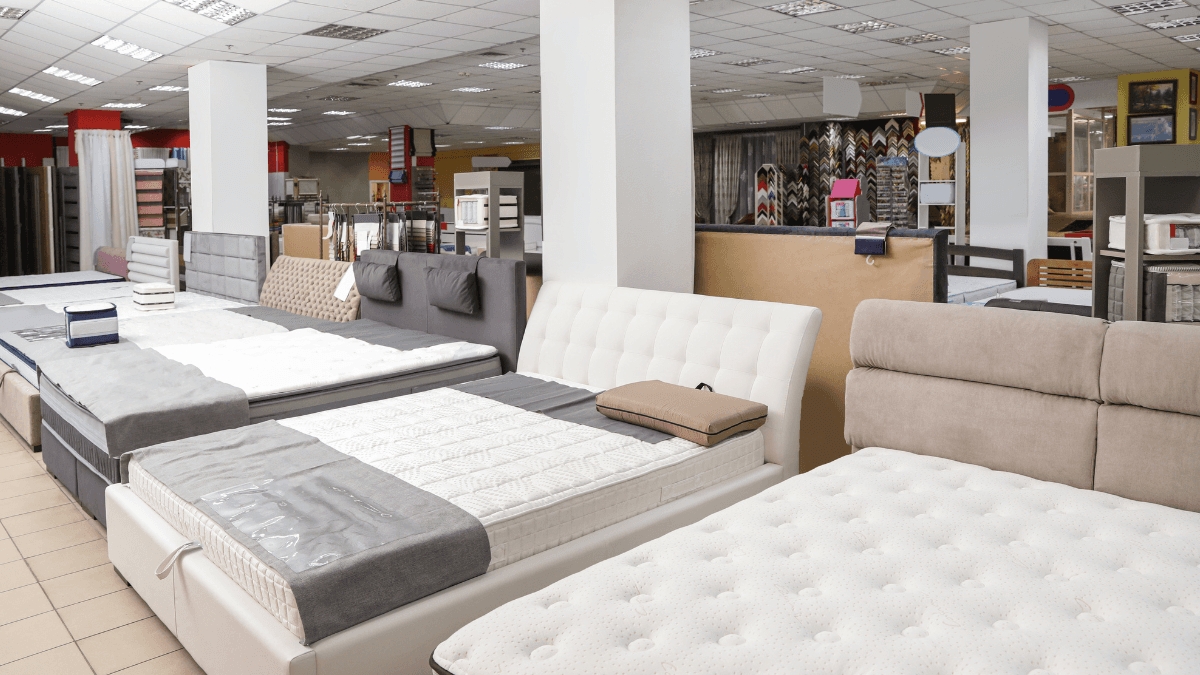 Interior of a mattress store showing various models of beds and mattresses on display, with different designs and colors arranged in a well-lit showroom.