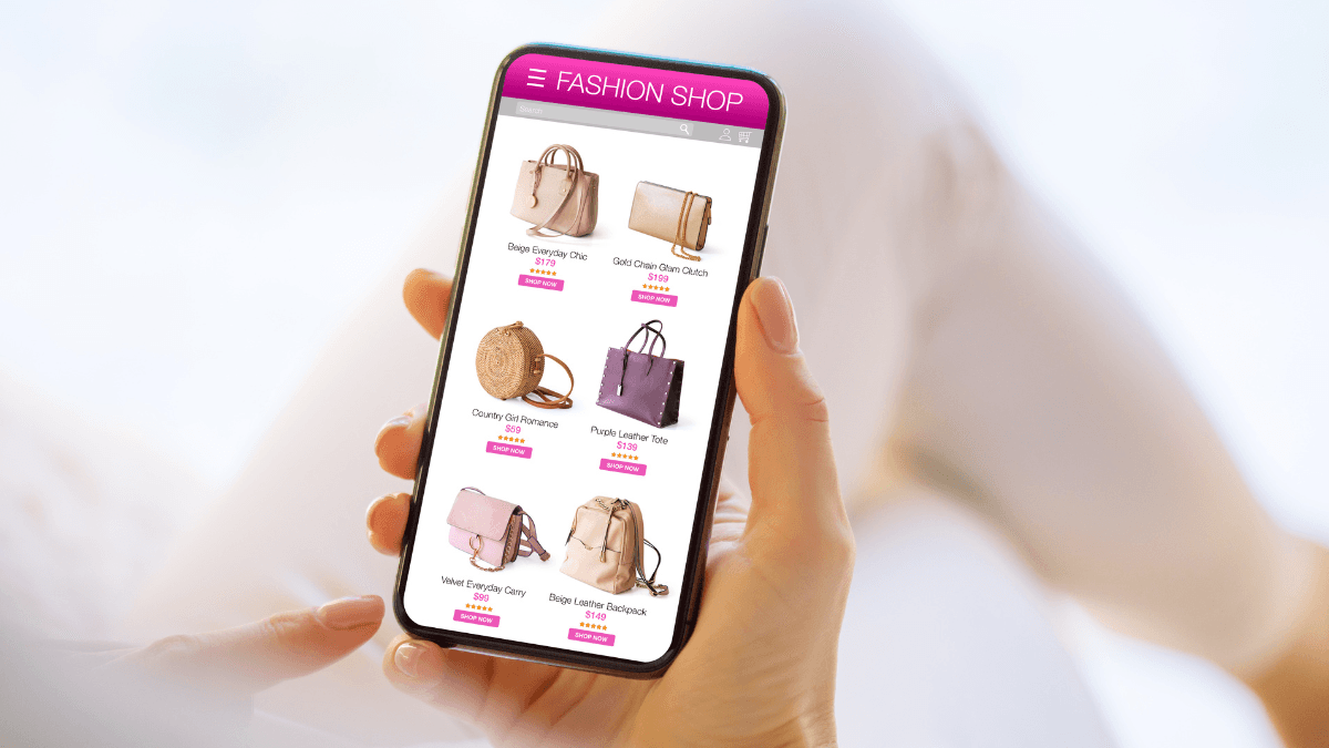 Person holding a smartphone displaying an online fashion shop with various handbags, featuring prices and 'shop now' buttons, set against a soft, blurred background for a focused look.