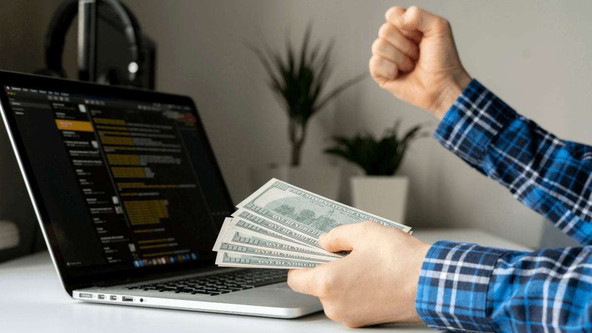 Close-up of a person holding dollar bills in front of a laptop screen displaying code or data. The individual's other hand is clenched in a fist, showing excitement or triumph, with a minimalistic workspace in the background.