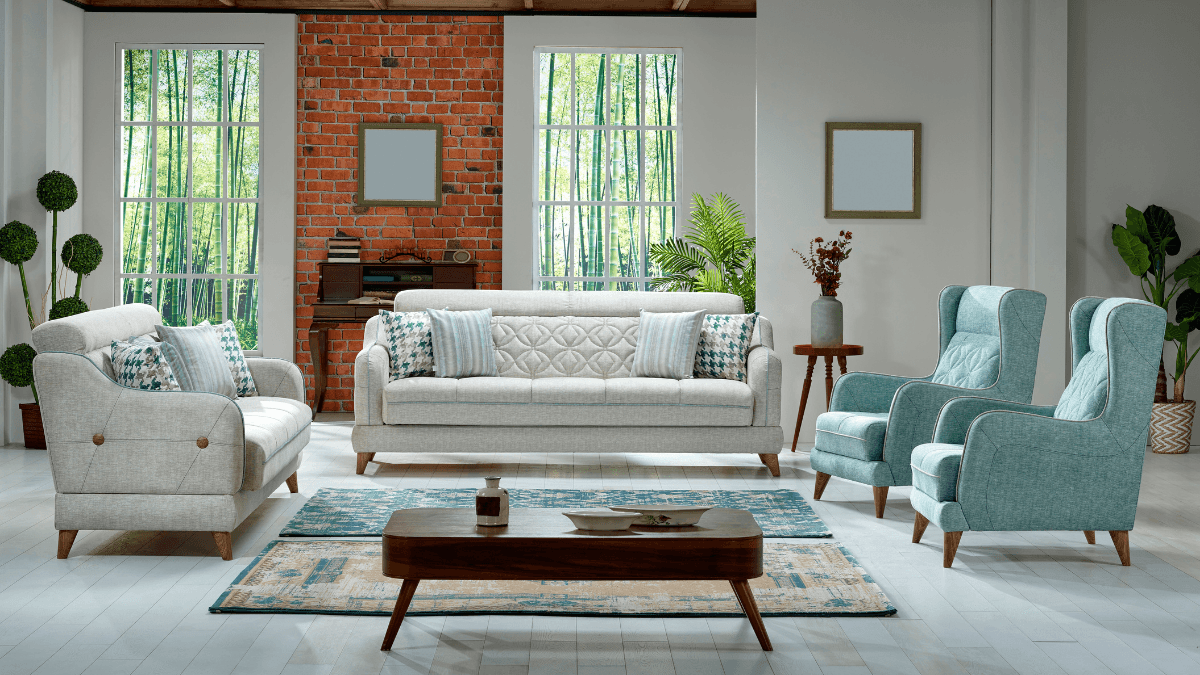 A cozy, modern living room setup featuring a combination of light and teal armchairs, a white sofa with accent pillows, and a rustic coffee table on a patterned rug, framed by large windows and greenery.