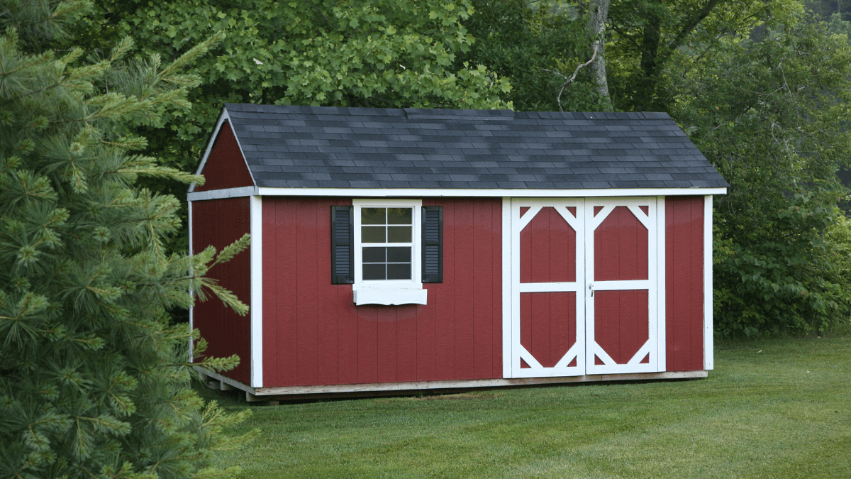 Classic red storage shed with white trim and a window, situated in a grassy yard and surrounded by trees, highlighting a rustic outdoor setting.