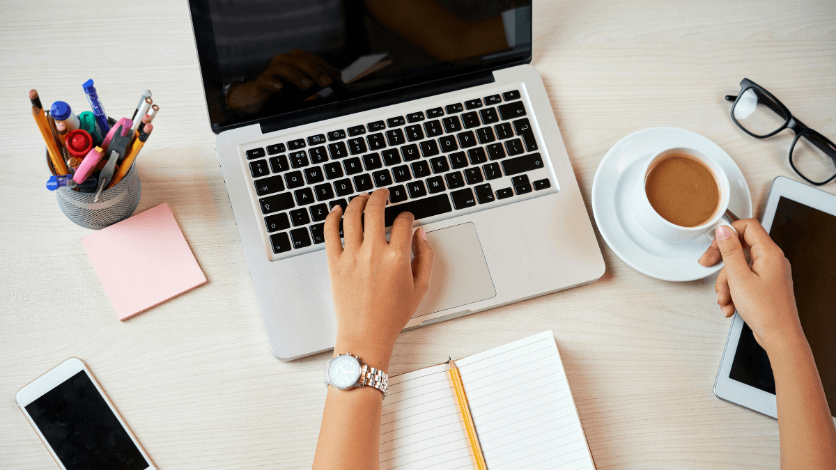 A workspace with a laptop, an open notebook with a pencil, a cup of coffee on a saucer, and various stationery items, including a pen holder, sticky notes, and a smartphone, all placed on a light wooden desk.